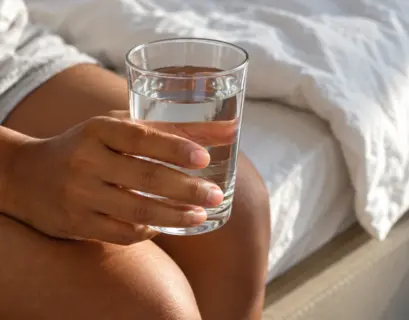 Mãos de uma pessoa segurando um copo de água transparente sentada na cama, com luz natural da manhã ao fundo.
