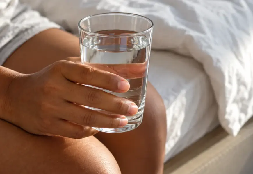 Mãos de uma pessoa segurando um copo de água transparente sentada na cama, com luz natural da manhã ao fundo.